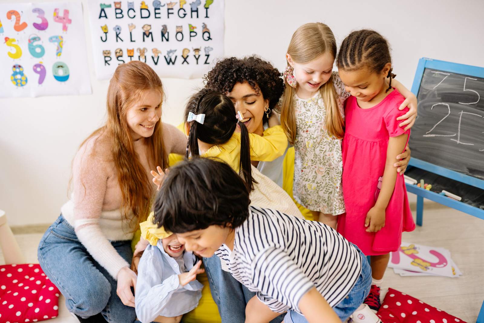 A preschool teacher kneeling with a small group of children in a bright classroom.
