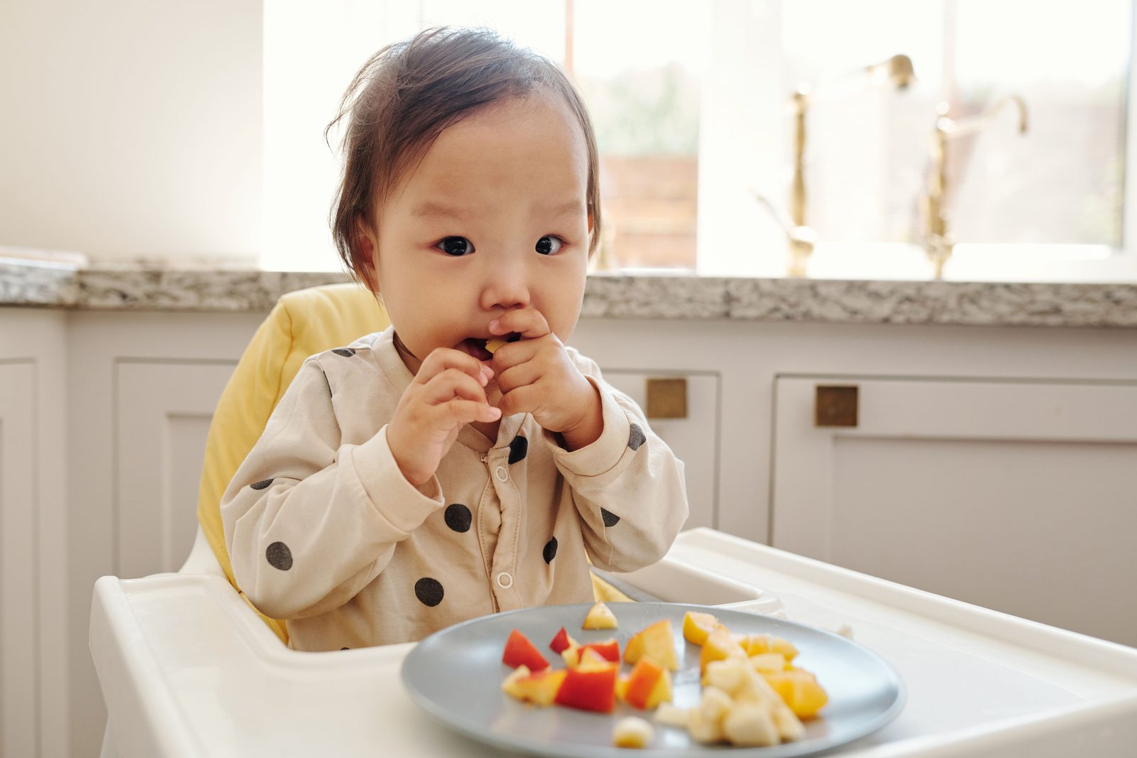 A toddler in a high chair eats pieces of fruit from a plate in a sunny kitchen.