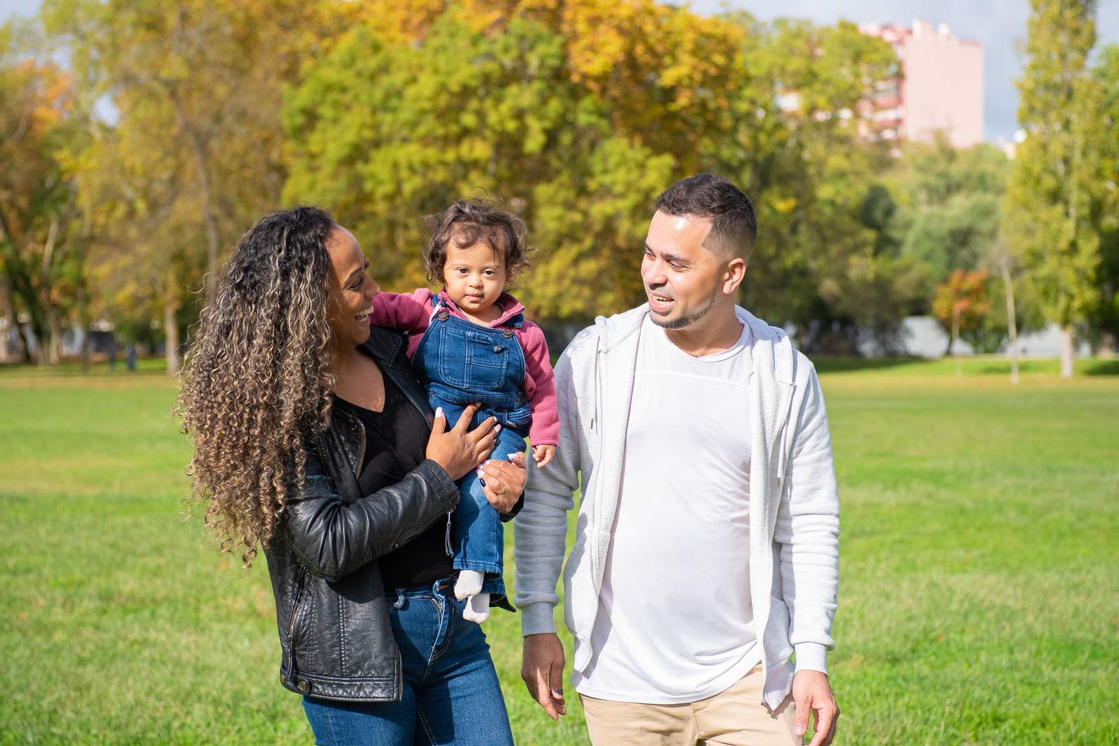 A mother and father walk in a park on a sunny day, carrying their toddler between them.