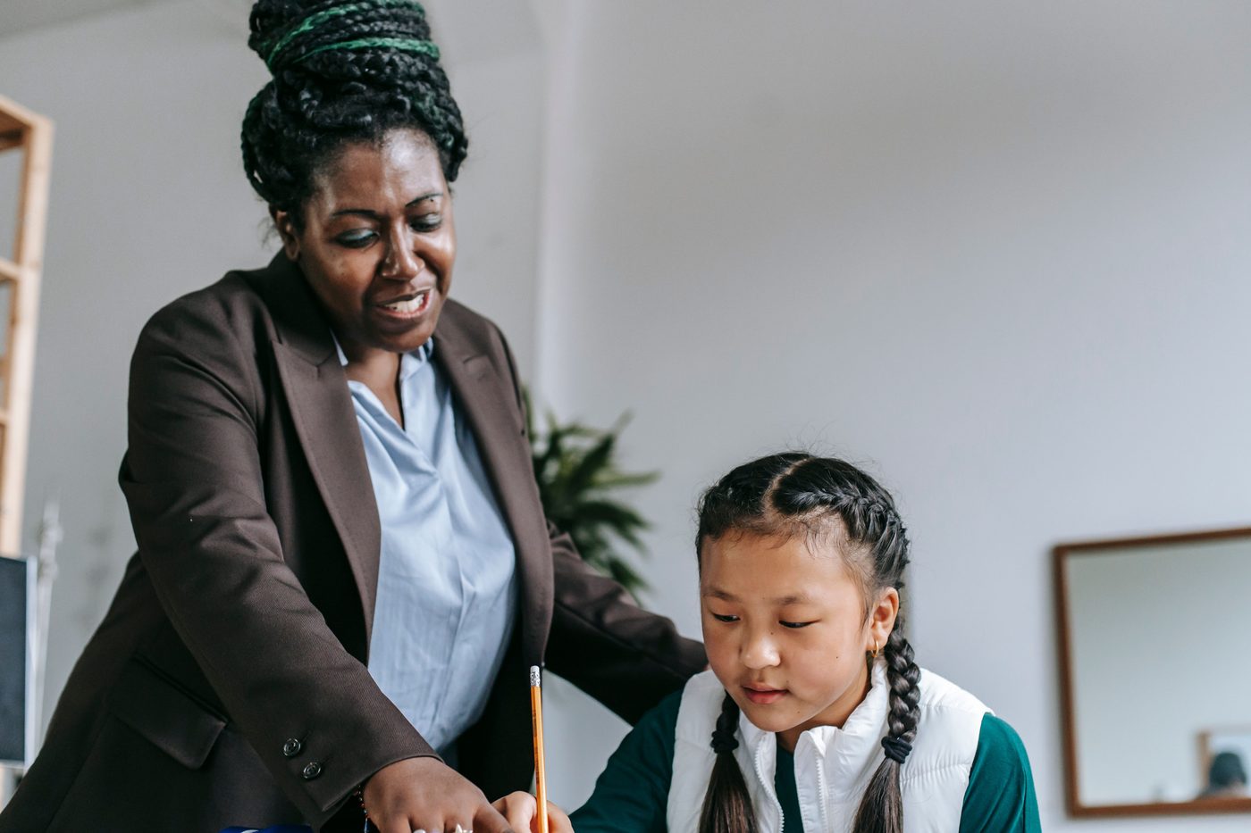 Middle aged African American female teacher with dreadlocks in formal clothes helping smart attentive little Asian girl 