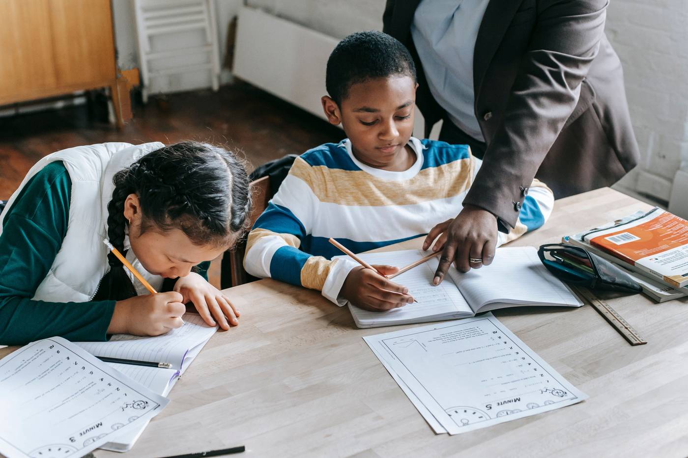 Black female teacher explaining task to focused little schoolboy sitting at desk near attentive Asian classmate writing 