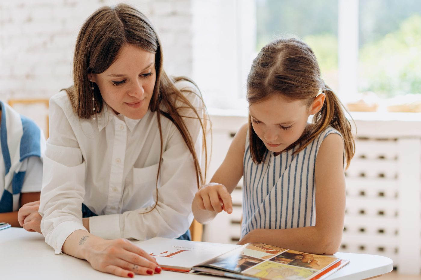 A teacher guides a young girl reading a book indoors