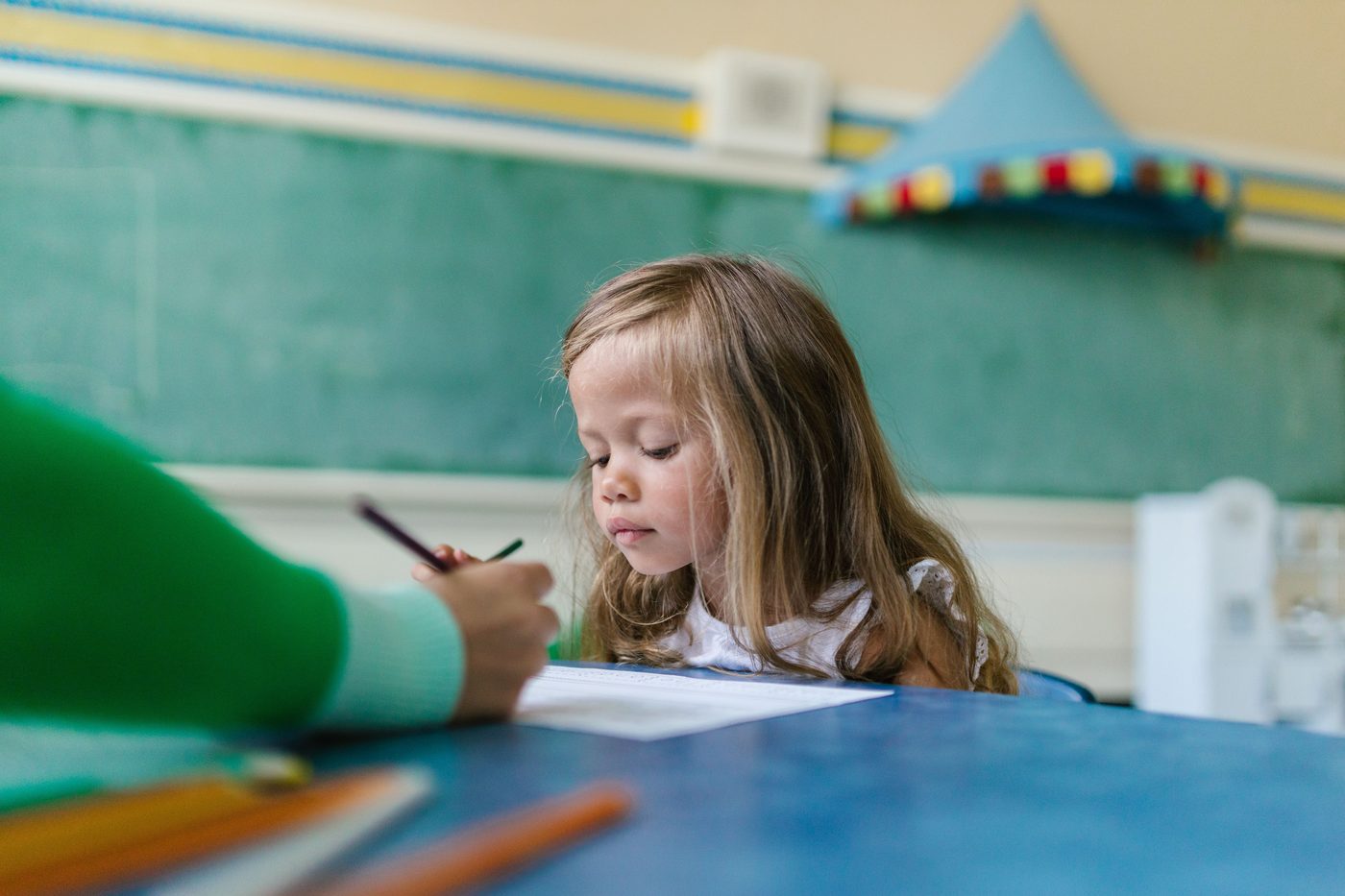 A young girl with long hair concentrates on her drawing in a colorful classroom environment