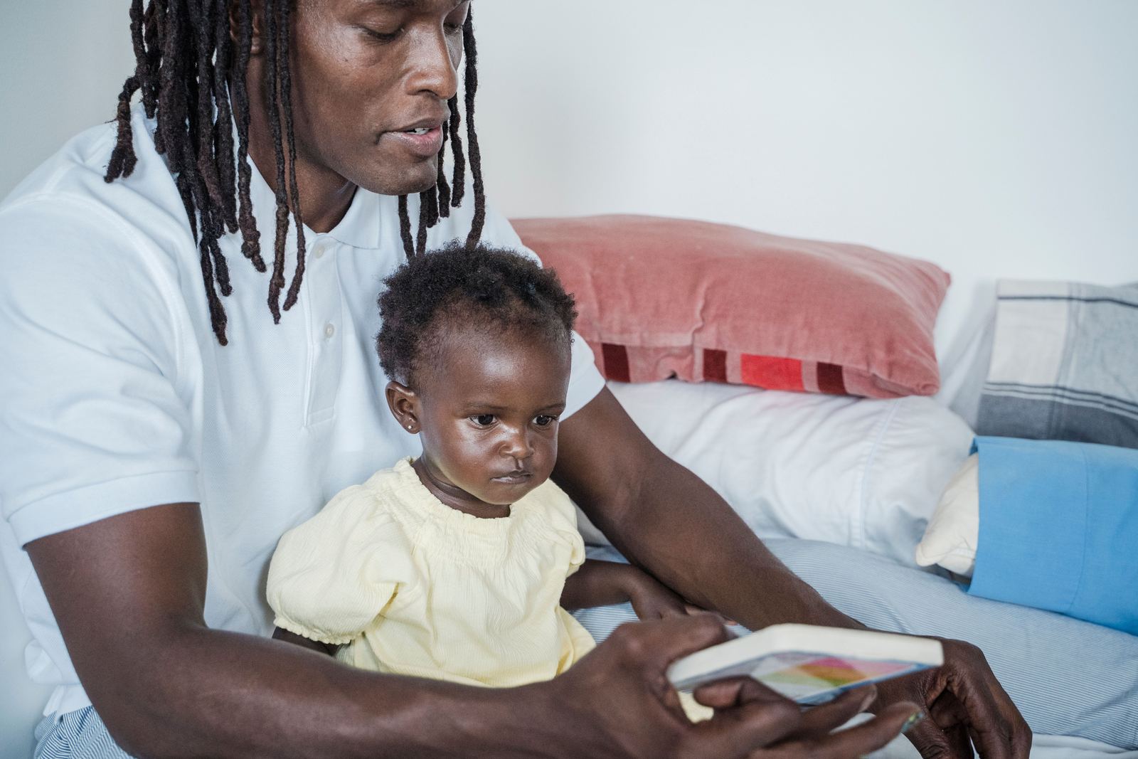 A father sits on the couch with his baby on his lap and reads a picture book with her.