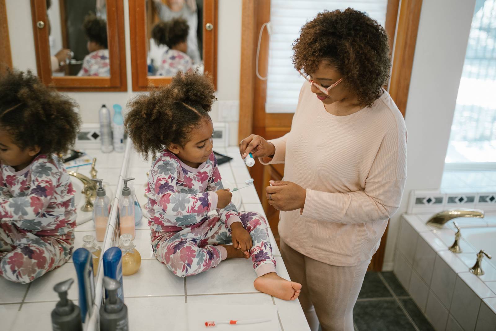 A parent helps a young child brush their teeth at a bathroom sink.
