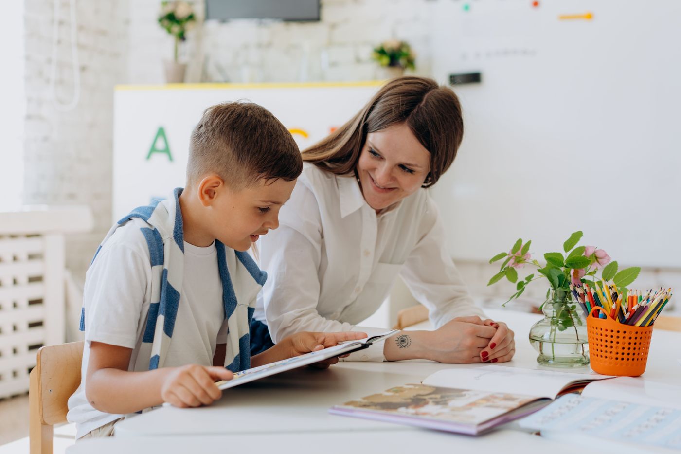 An adult sits beside a boy using a tablet at a classroom table