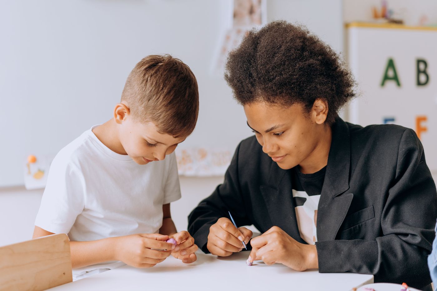 An adult and a boy work side by side on a writing task at a classroom desk