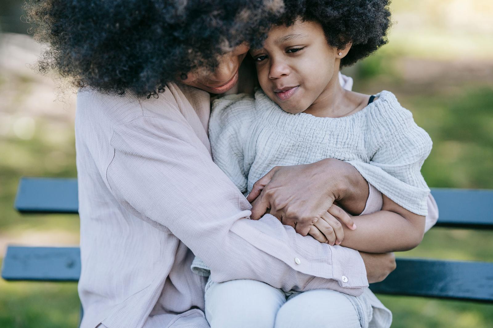A mother kneels on a park bench and hugs her young daughter close after a hard moment.
